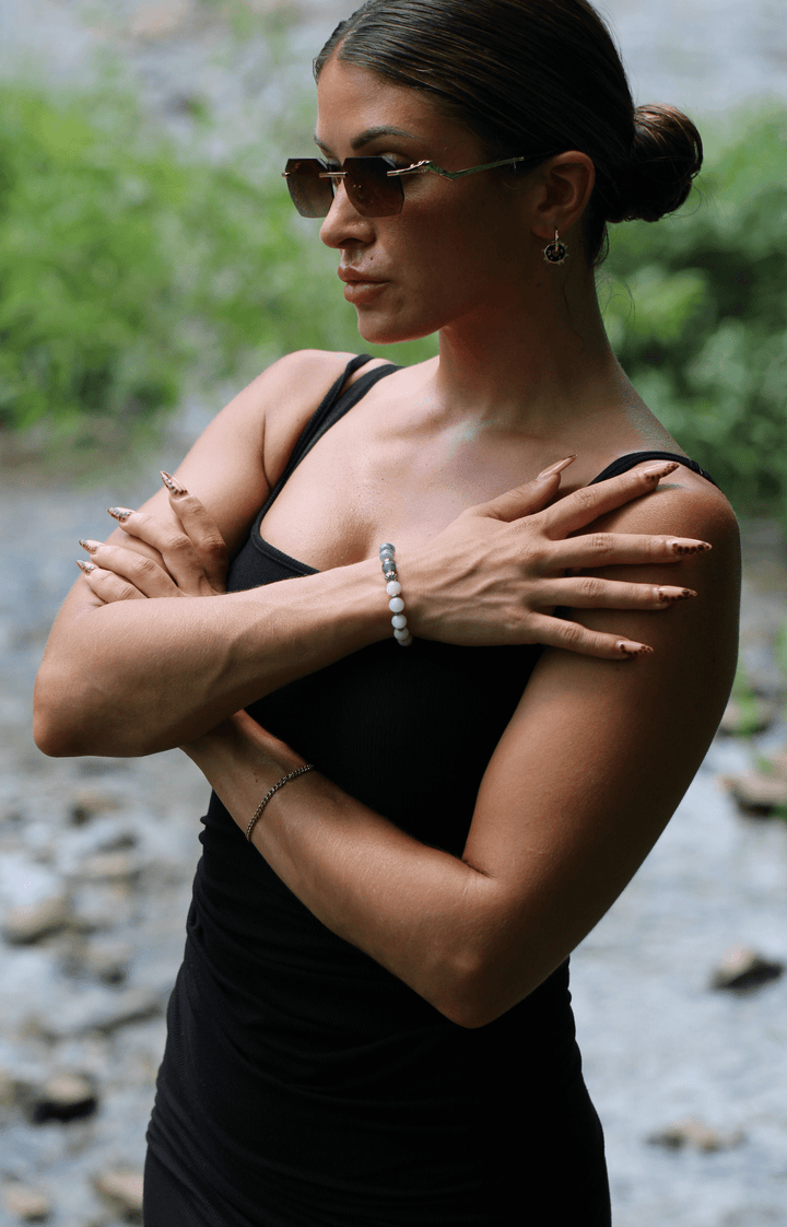 Model wearing the Teach kunzite and amethyst bracelet with arms crossed beside a stream