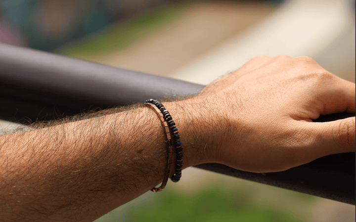 Titanium Hematite Rectangle bracelet stacked with dark beads on a wrist by an outdoor railing